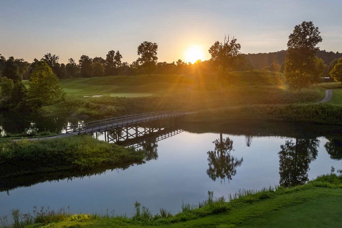 A sun sets over a serene landscape featuring a bridge over a reflective body of water, surrounded by lush greenery and trees.