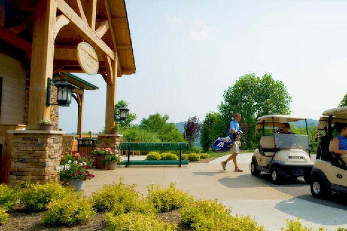 A person carrying golf clubs near a clubhouse, with two golf carts parked nearby and lush greenery surrounding the area.