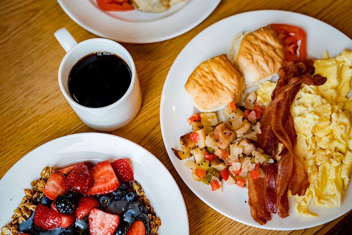 The image shows a breakfast spread with scrambled eggs, bacon, biscuits, home fries, a bowl of mixed berries, and a cup of coffee on a wooden table.