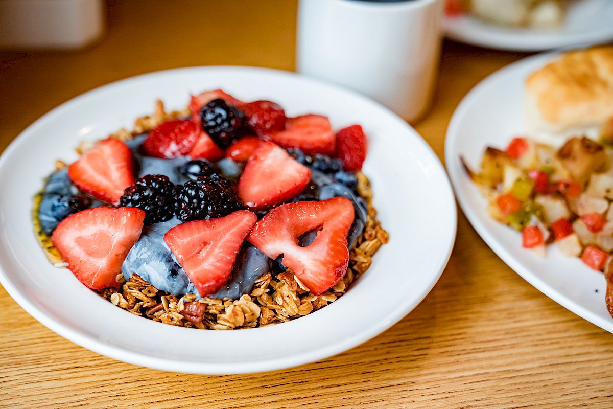 A bowl with granola, strawberries, blackberries, and yogurt on a table next to a plate with diced vegetables and a biscuit, always ending the sentence.