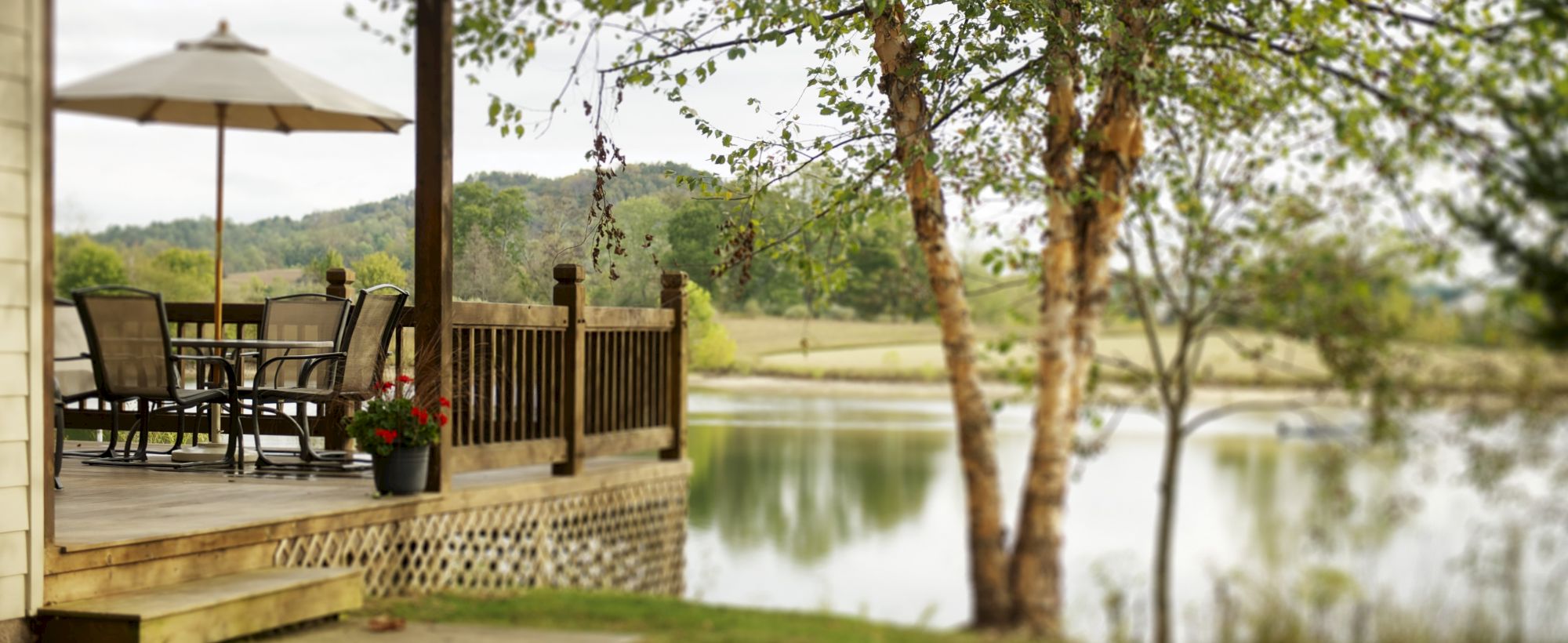 A cozy porch overlooking a peaceful lake with outdoor furniture, an umbrella, and a potted plant, surrounded by trees and serene landscape views.