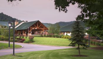 A landscaped area with a paved road, lush green grass, a tree, and a building with a green roof set against a backdrop of hills and trees, ends the sentence.
