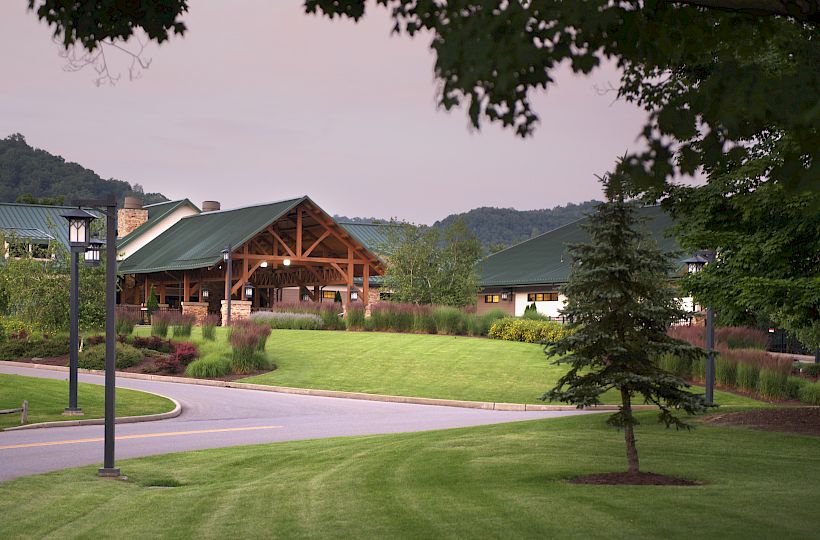 A landscaped area with a paved road, lush green grass, a tree, and a building with a green roof set against a backdrop of hills and trees, ends the sentence.