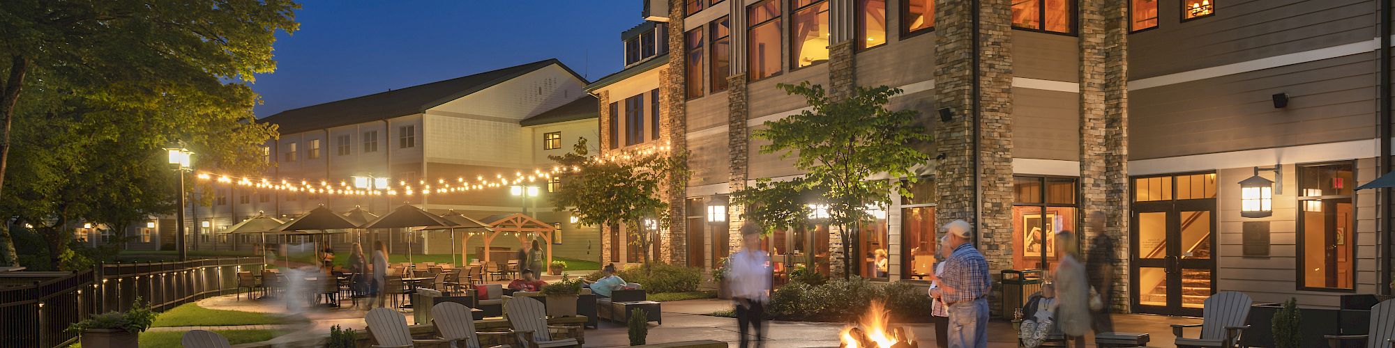 The image shows a nighttime scene of a large, well-lit building with an outdoor seating area around a fire pit, with people socializing.